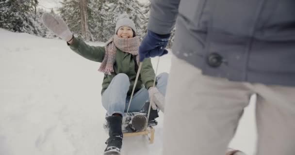 Медленный портрет счастливой азиатки, катающейся на санках со смехом разговаривающей с парнем в зимнем лесу — стоковое видео