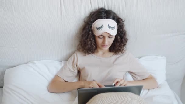 ベッドの中でラップトップを使用してパジャマを身に着けている楽しい女の子は、オンライン通信を楽しんで入力笑顔. — ストック動画