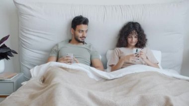 Arap bir adam ve beyaz bir kız akıllı telefonlar kullanıyor. Yatakta birlikte uzanıyorlar. Boş zamanlarında evde dinleniyorlar.