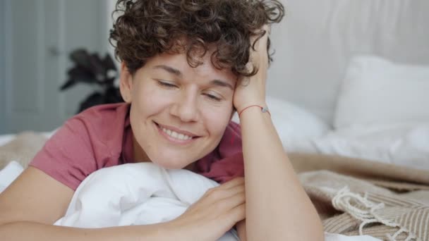 Retrato de bela mulher de cabelos encaracolados sorrindo e tocando o cabelo deitado na cama em casa — Vídeo de Stock