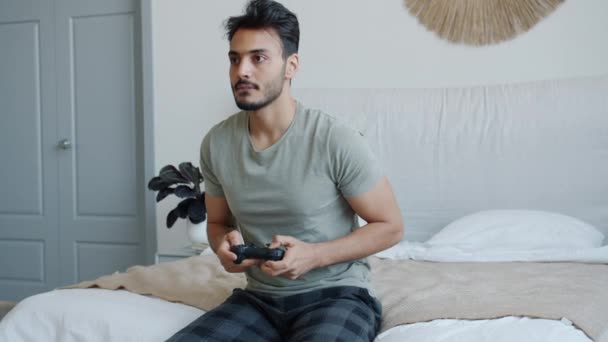 Emocional hombre de Oriente Medio jugando videojuego gritando divirtiéndose sentado en la cama en casa — Vídeos de Stock