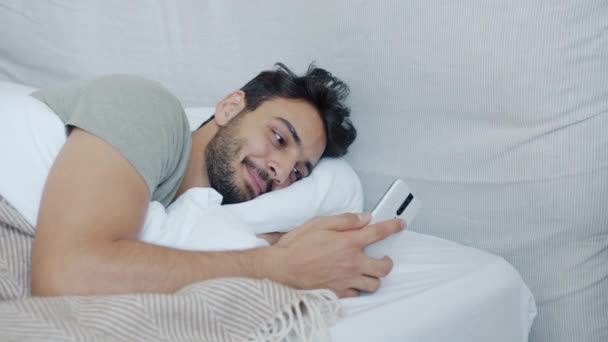 Glada Mellanöstern kille som ligger i sängen och använder smartphone pekskärm leende — Stockvideo