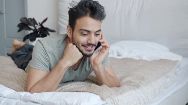 Hombre árabe joven charlando en el teléfono móvil en la cama en el apartamento disfrutando de la conversación — Vídeo de stock