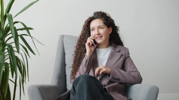 Retrato de una joven y ambiciosa mujer de negocios hablando por teléfono móvil y sonriendo sentada en un sillón — Vídeo de stock