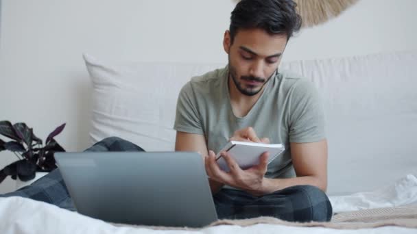 Hombre árabe ambicioso freelancer que trabaja en línea usando el ordenador portátil y la escritura en el cuaderno en la cama — Vídeos de Stock