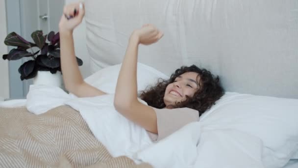 Радостная девушка танцует лежа в постели наслаждаясь беззаботным утром дома — стоковое видео