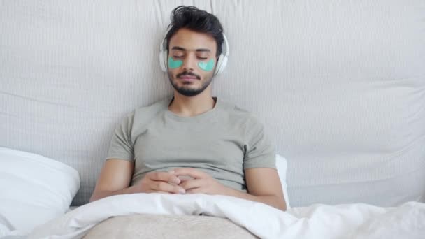Bliskowschodni mężczyzna z przepaskami na oczy słuchający muzyki ze słuchawkami i tańczący w łóżku — Wideo stockowe