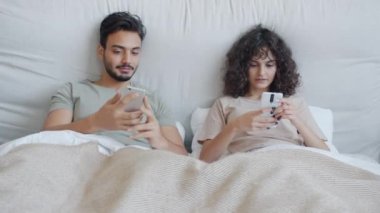 Mutlu karışık ırk çifti akıllı telefon kullanıyor, konuşuyor ve gülüyor evde yatakta dinleniyor.