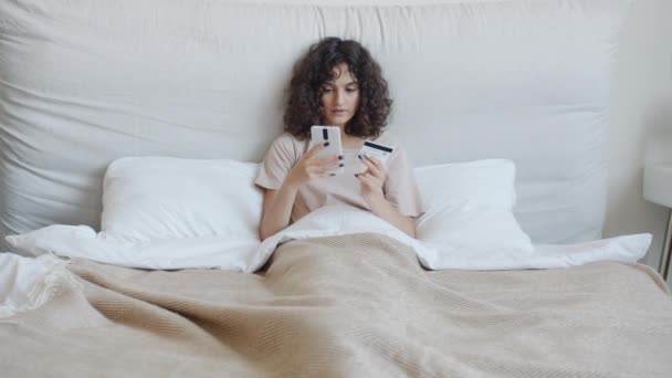 Chica alegre de compras en línea con teléfono inteligente que paga con tarjeta bancaria sonriendo en la cama — Vídeo de stock