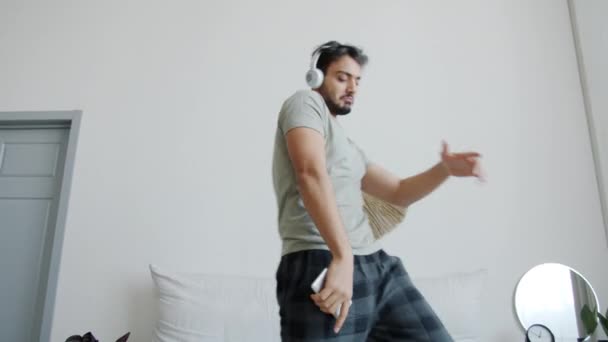 Portrait of happy Arab guy dancing in bed wearing headphones and holding smartphone — Stock Video