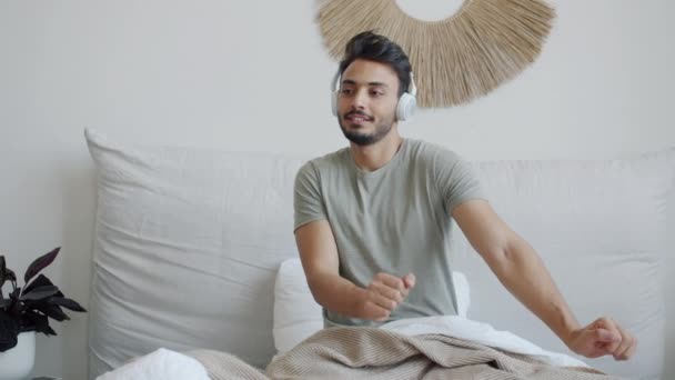 快乐的阿拉伯小伙子在床上跳舞，通过耳机听音乐，在家里玩得很开心 — 图库视频影像