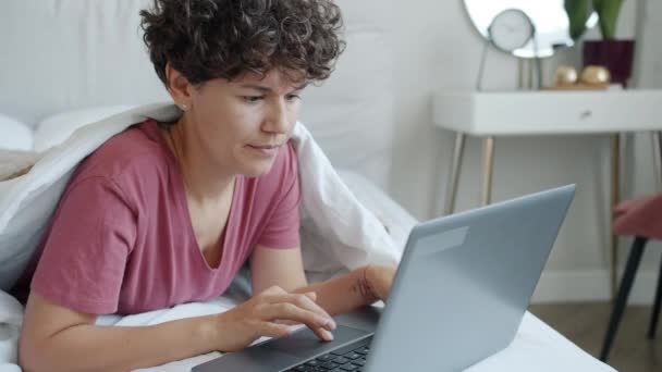 Joyeux jeune dame aux cheveux bouclés en utilisant un ordinateur portable couché dans le lit à la maison — Video