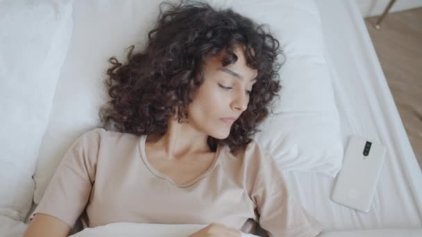 Visão superior câmera lenta da menina de cabelos encaracolados acordar na cama tocando tela do smartphone e braços esticados — Vídeo de Stock