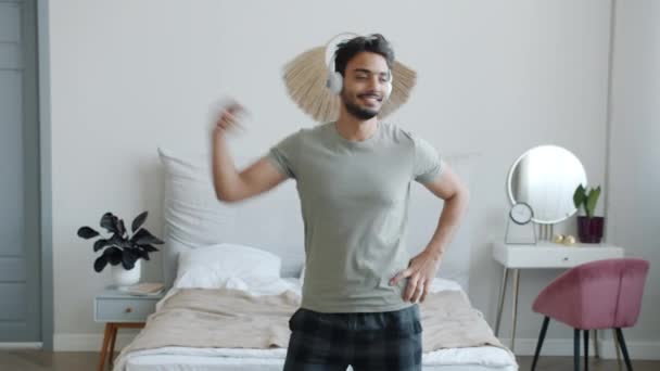 无忧无虑的阿拉伯年轻人戴着耳机，手持智能手机，在卧室里跳舞 — 图库视频影像