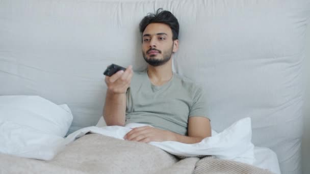Парень с Ближнего Востока смотрит телевизор и смеется, расслабляясь в постели дома — стоковое видео
