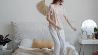Pijama giyen, dans eden ve yatakta zıplayan mutlu genç bayan evde eğleniyor.