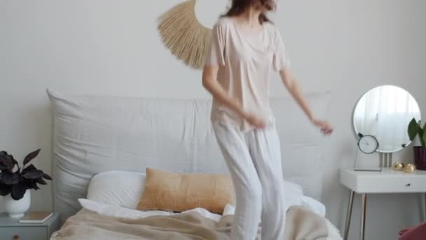 Glad ung dam i pyjamas dansar och hoppar på sängen ha kul hemma — Stockvideo