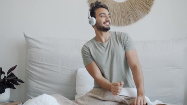 Zeitlupe: Unbekümmerter Student aus dem Nahen Osten tanzt im Bett und genießt Musik über Kopfhörer — Stockvideo