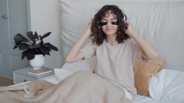 Menina feliz com manchas nos olhos ouvindo música através de fones de ouvido dançando na cama — Vídeo de Stock