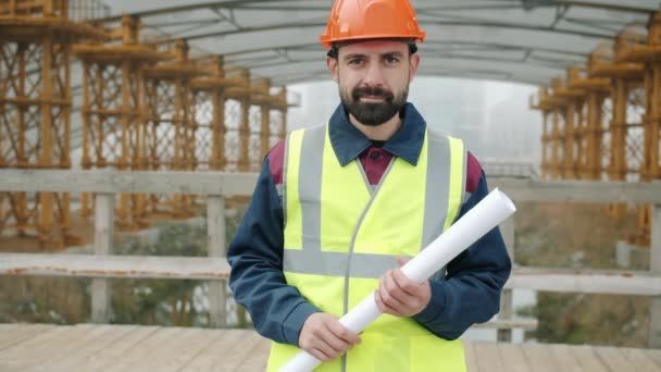 Retrato del constructor masculino con casco y chaleco parado fuera del sitio de construcción sosteniendo el plano — Vídeo de stock