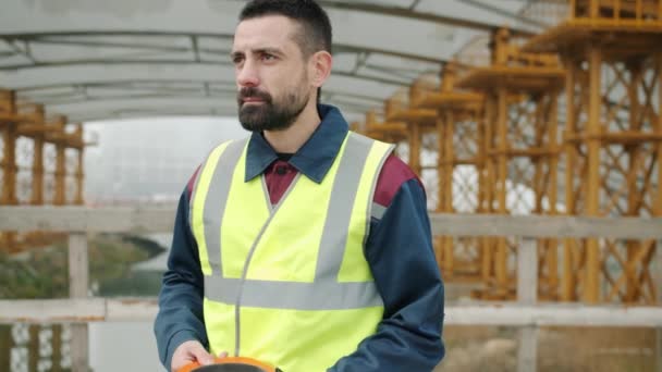 Trabajador de la construcción que se pone casco de seguridad y cruza brazos de pie en la zona del edificio — Vídeos de Stock