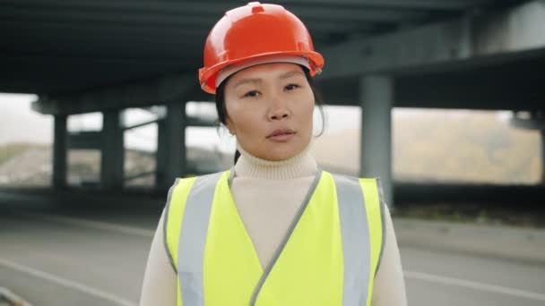Портрет серьезной азиатки в строительной форме, стоящей только в строительной зоне — стоковое видео