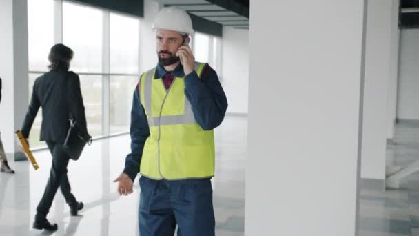 Powolny portret inżyniera noszącego kask i kamizelkę mówiącego przez telefon komórkowy w nowym budynku — Wideo stockowe