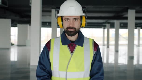 Retrato en cámara lenta del constructor que usa ropa de seguridad y auriculares de pie en un edificio comercial — Vídeo de stock
