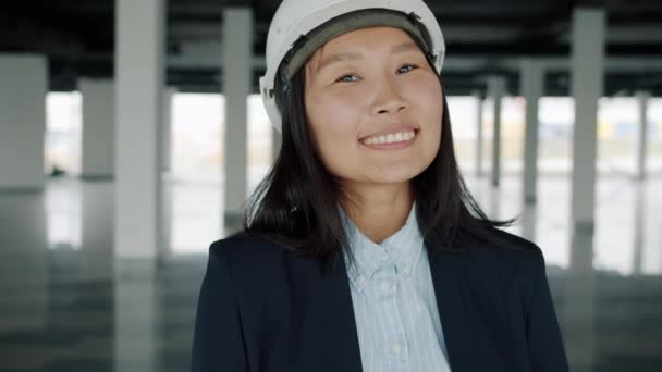 Retrato de mujer asiática ingeniera de pie en el interior de un nuevo edificio con casco de seguridad sonriendo — Vídeos de Stock