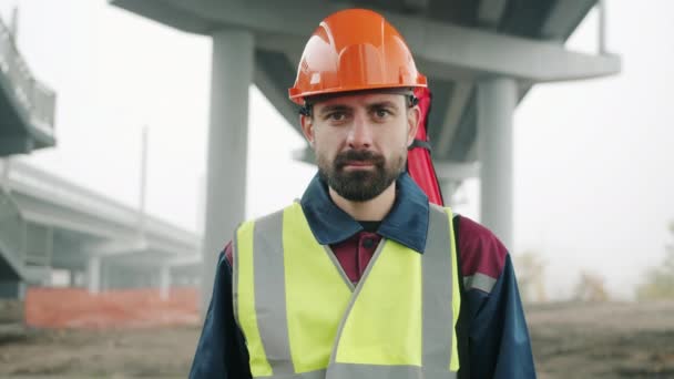 Портрет геодезиста в шоломі безпеки і жилеті, що стоїть на будівельному майданчику з геодезичним обладнанням на задньому плані — стокове відео