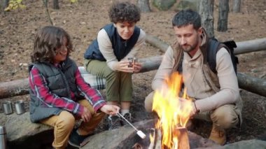 Küçük çocuk şekerleme pişiriyor ve dışarıda kamp ateşinin tadını çıkaran ebeveynlerle konuşuyor.