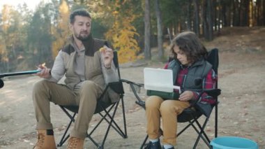 Küçük çocuk, nehir kıyısındaki sandalyede oturan babasına balık yemi veriyor.