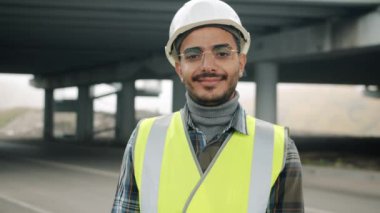 İnşaat üniforması giyen neşeli bir Arap inşaat alanında durup gülümsüyor.