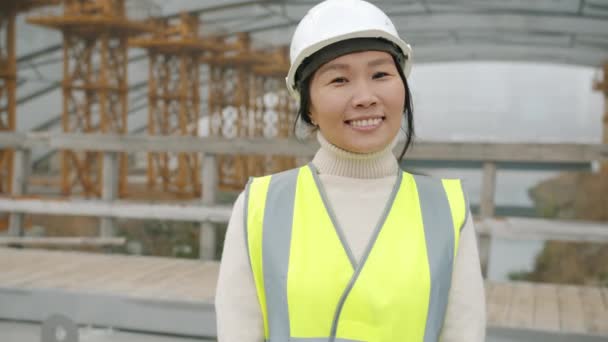 Портрет веселой азиатской леди в шлеме и защитном жилете, улыбающейся в зоне строительства — стоковое видео