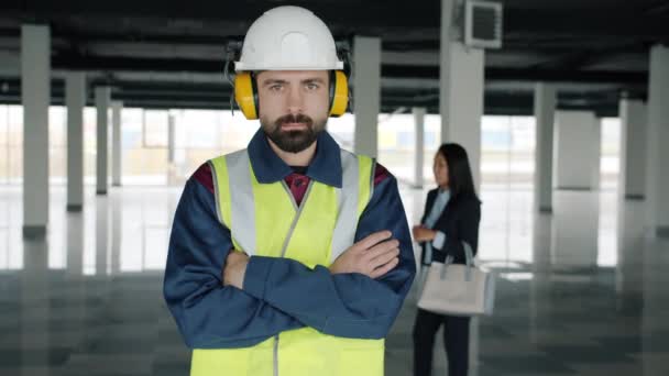 Портрет специалиста по обслуживанию, стоящего внутри нового промышленного здания, в то время как бизнесмены разговаривают на заднем плане — стоковое видео