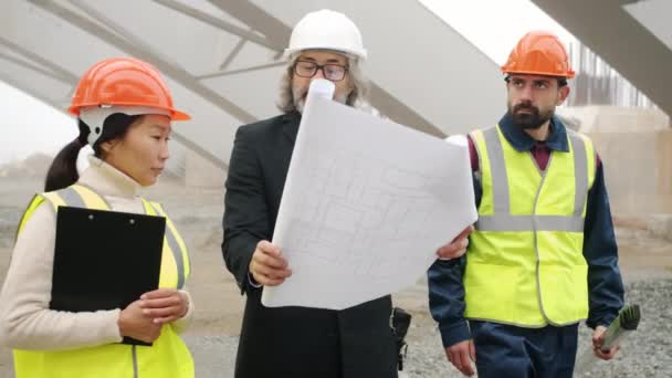 Arquitecto hablando con trabajadores de la construcción mostrando planos caminando en la zona de edificios al aire libre — Vídeo de stock