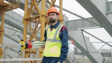 İnşaat alanında tek başına yürüyen teodolit taşıyan ciddi bir adamın Dolly çekimi.