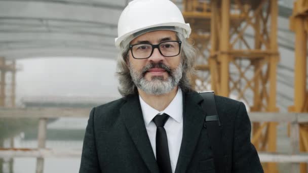 Retrato en cámara lenta del exitoso arquitecto hombre maduro que usa casco de seguridad de pie en el área de construcción — Vídeo de stock