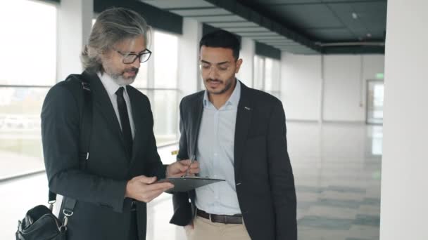 Bliskowschodni biznesmen rozmawiający z partnerem kaukaskim podpisuje dokument w nowoczesnym budynku przemysłowym — Wideo stockowe