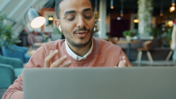 Retrato de un árabe feliz charlando en una videollamada en un café con vainas de aire y usando un portátil mientras la gente camina en el fondo — Vídeo de stock