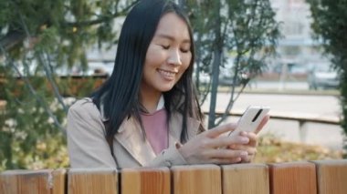 Dışarıdaki akıllı telefon ekranına bakıp gülümseyen Asyalı kadının yavaş çekim portresi.