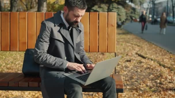 Movimento lento de homem de negócios sério usando laptop sentado no banco ao ar livre — Vídeo de Stock