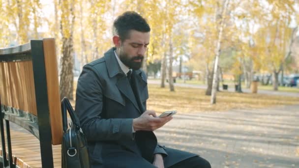 Retrato del joven empresario hablando por teléfono móvil sentado en el parque de la ciudad en el día de otoño — Vídeo de stock