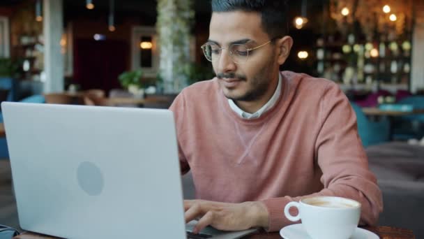 Retrato en cámara lenta de un joven árabe usando un ordenador portátil y tomando café en una bonita cafetería — Vídeo de stock