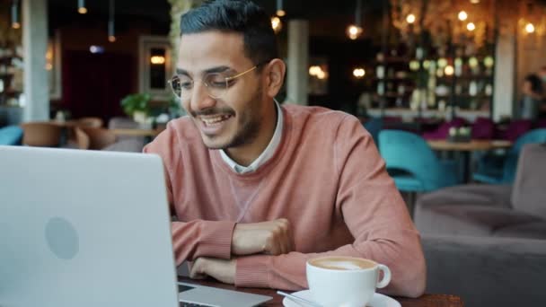 Retrato del joven y feliz hombre de Oriente Medio charlando en videollamada y saludando con la mano usando el ordenador portátil en la cafetería — Vídeo de stock