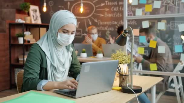 Muslim businesswoman wearing face mask using laptop then talking to male colleague in office — Stock Video