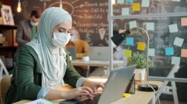 Başörtüsü ve yüz maskesi takmış Müslüman bir iş kadını dizüstü bilgisayar kullanıp ofiste cep telefonu görüşmesi yapıyor.