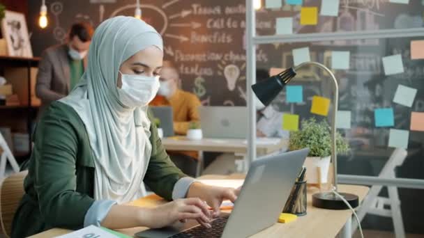 Başörtüsü ve yüz maskesi takmış Müslüman bir iş kadını dizüstü bilgisayar kullanıp ofiste cep telefonu görüşmesi yapıyor. — Stok video