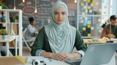 Ofiste masa başında oturan ve ciddi bir yüzle kameraya bakan tesettürlü müslüman bir ofis çalışanının yavaş çekim portresi. İnsan ve iş kavramı.