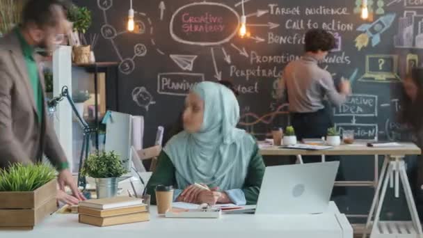 El lapso de tiempo del empleado musulmán en el hijab trabajando con el ordenador portátil y discutiendo negocios con colegas en la oficina creativa — Vídeo de stock